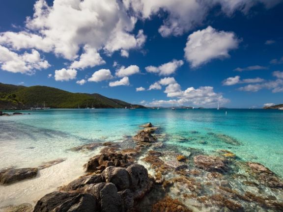 View of Francis Beach in the US Virgin Islands (USVI), from the beach with rocks in the foreground, overlooking the crystal-clear turquoise water with yachts on the horizon.